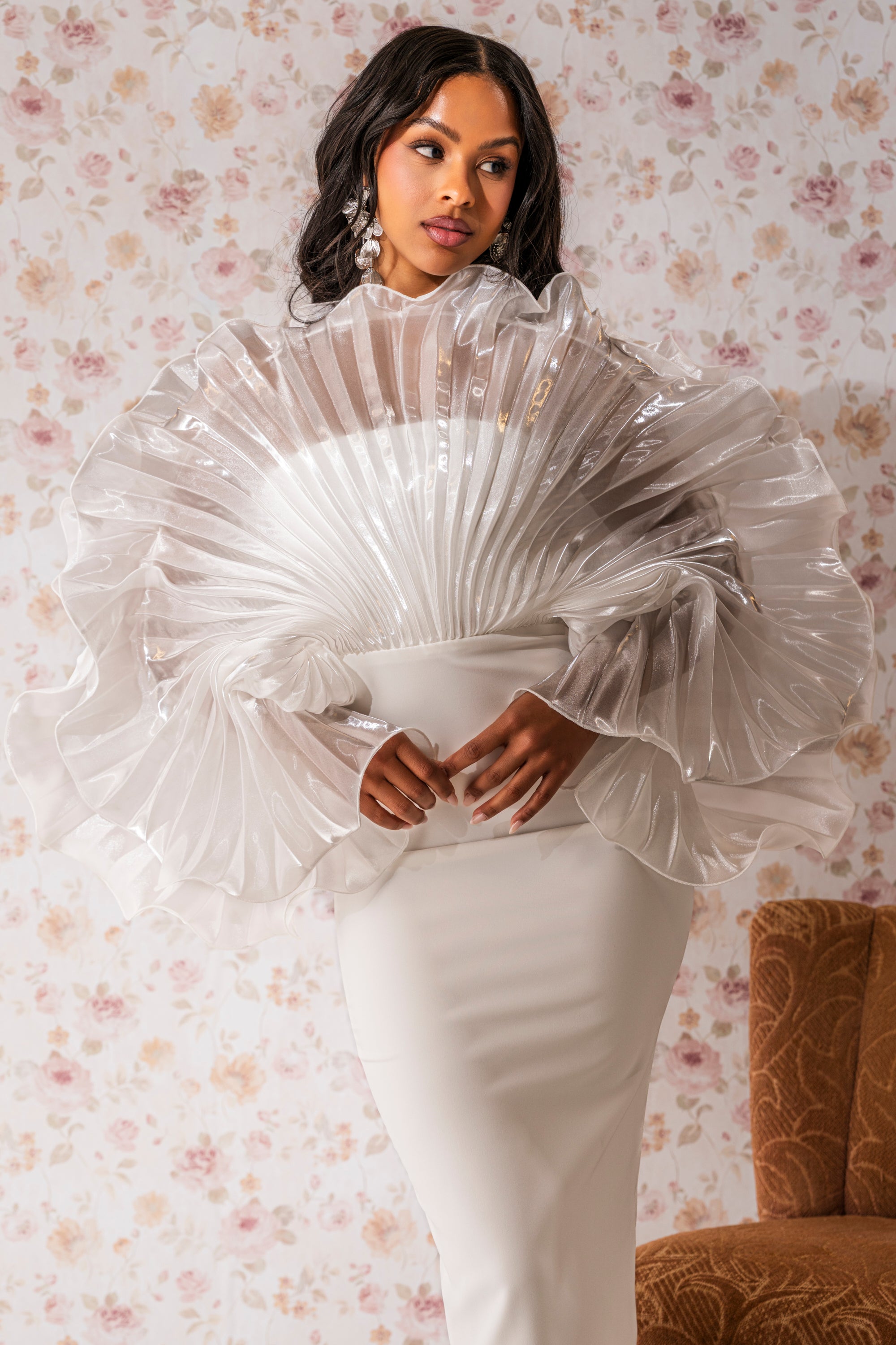 A woman stands before floral wallpaper, wearing the BY THE SEA SHORE BANDAGE MIDI DRESS featuring a dramatic sheer, fan-pleated bodice and voluminous sleeves. Her hair is in loose waves and she accessorizes with statement earrings.