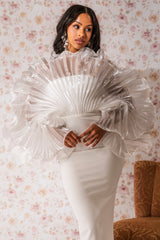 A woman stands before floral wallpaper, wearing the BY THE SEA SHORE BANDAGE MIDI DRESS featuring a dramatic sheer, fan-pleated bodice and voluminous sleeves. Her hair is in loose waves and she accessorizes with statement earrings.