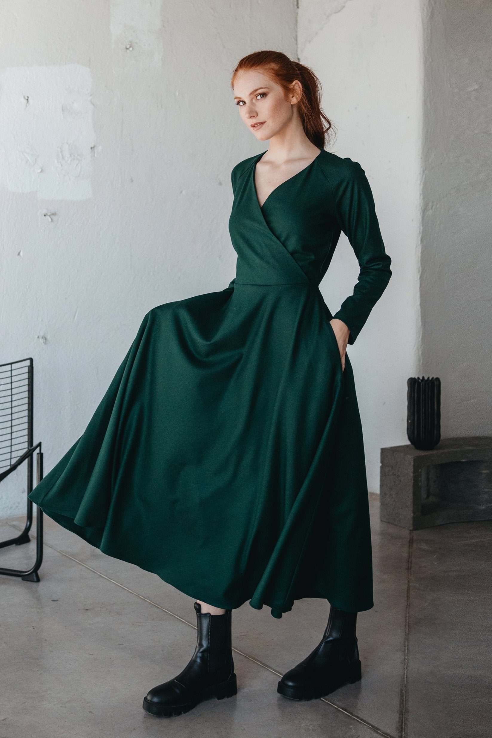 Woman wearing a deep green wool wrap-top dress with long sleeves and pockets, paired with black boots, standing indoors
