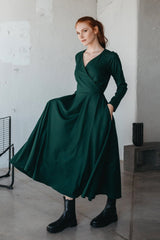 Woman wearing a deep green wool wrap-top dress with long sleeves and pockets, paired with black boots, standing indoors