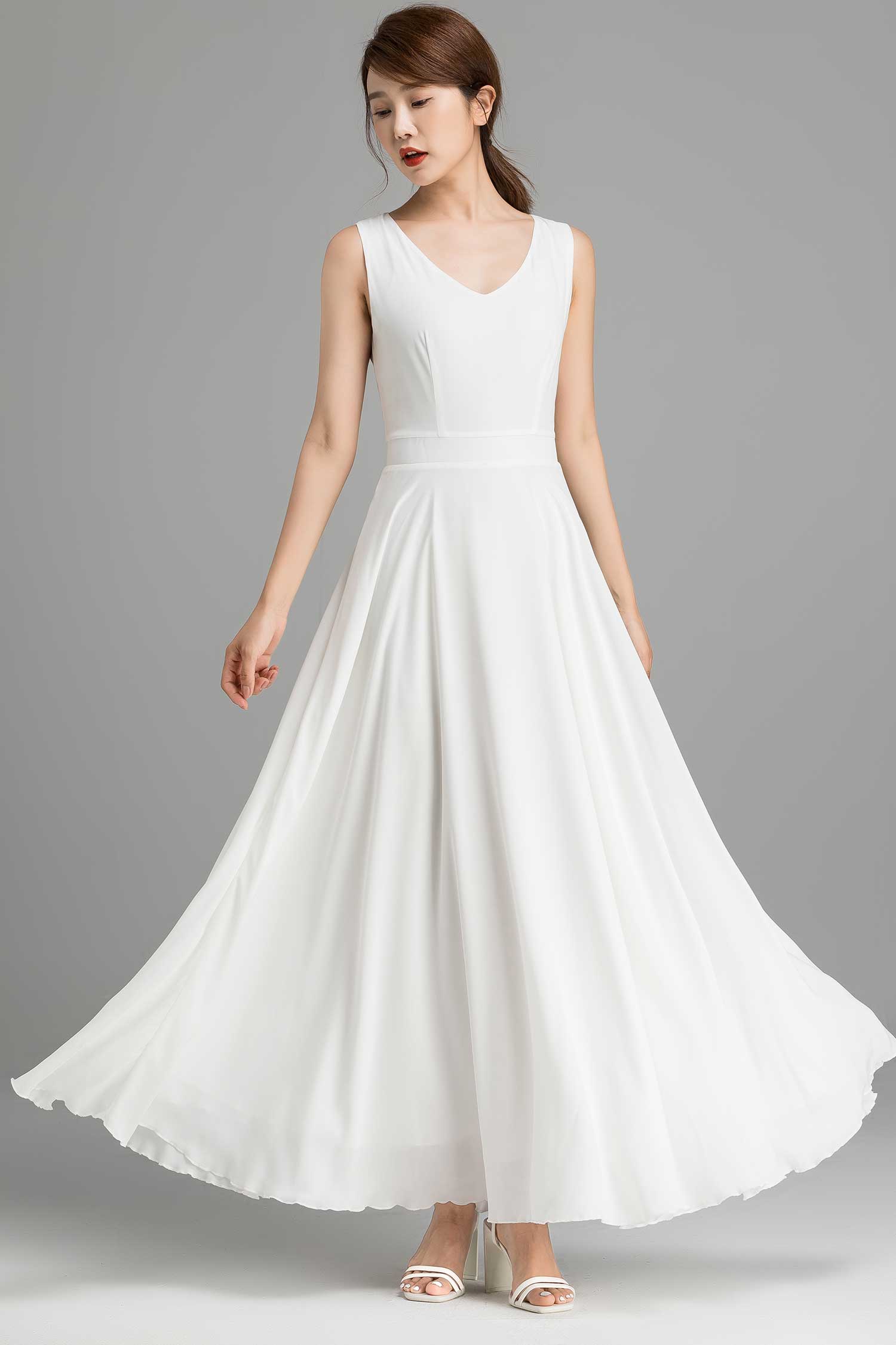 A woman modeling a white chiffon maxi dress with a V neckline and back zip closure, standing on a plain background.