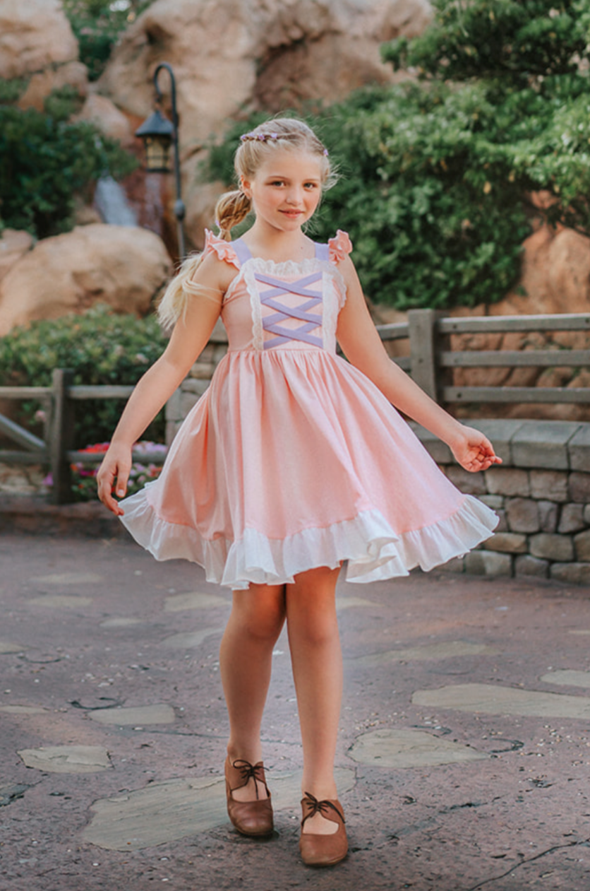 A young girl wearing Our Original Rapunzel Twirl Dress with a lavender laced bodice and white frills twirls outdoors. The dress is a light pink with white ruffle at the hem.  The sleeveless dress has light pink ruffles on lavender straps.