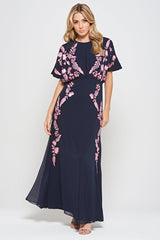 Navy floral embroidered maxi dress with pink detailing, worn by a model. Features flutter sleeves, a fitted waist and a flowing full-length skirt—perfect for weddings, evening occasions and special events.