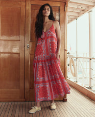 A woman stands on a wooden deck in THE GREAT. SU26's The Drop Waist Dress – Red Sailboat Bandana, paired with beige shoes. A wooden door and lifebuoy are visible in the background.