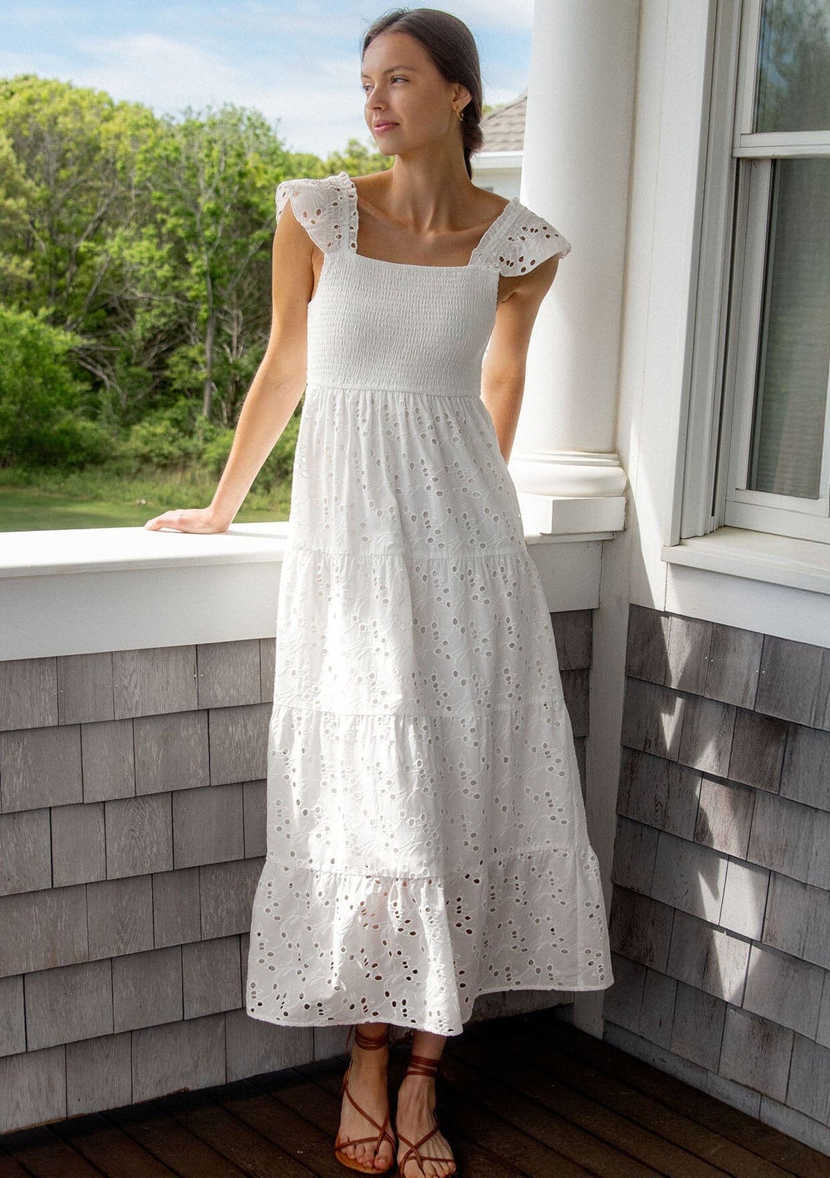 [Color: White] A front facing image of a brunette model wearing a bohemian cotton maxi dress in a teal embroidered eyelet. With a slim fit smocked bodice, a square neckline, short flutter sleeves, and a flowy long tiered skirt.