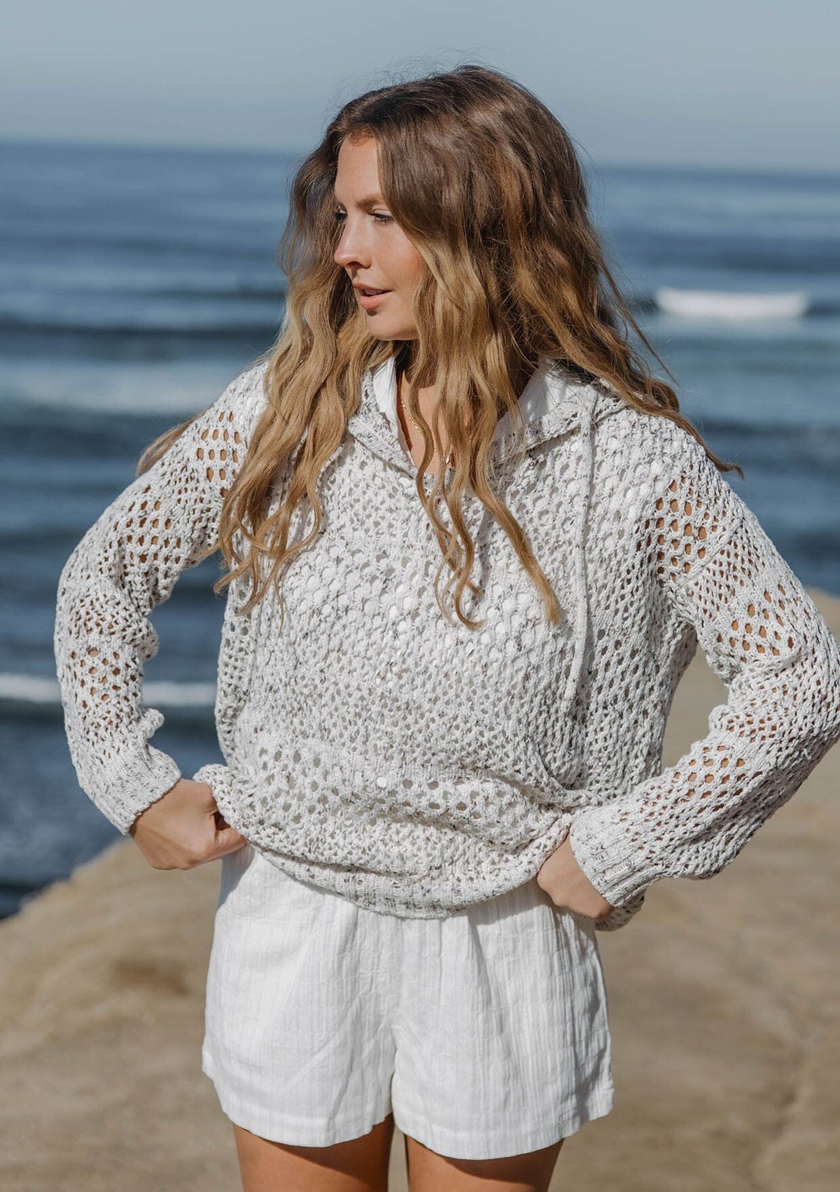 [Color: Ivory/Taupe] A front facing image of a blonde model wearing a lightweight crochet hoodie in an ivory and taupe speckled knit. With long sleeves, a v neckline, a drawstring hoodie, and a dropped shoulder.