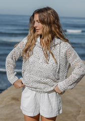 [Color: Ivory/Taupe] A front facing image of a blonde model wearing a lightweight crochet hoodie in an ivory and taupe speckled knit. With long sleeves, a v neckline, a drawstring hoodie, and a dropped shoulder.