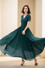 A woman in a flowing teal chiffon maxi dress with a V-neckline and short sleeves, dancing with her hands raised and flowing skirt.