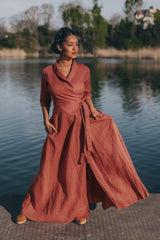 Woman wearing a rust-colored linen A-line dress with tie waist and V-neck standing by water