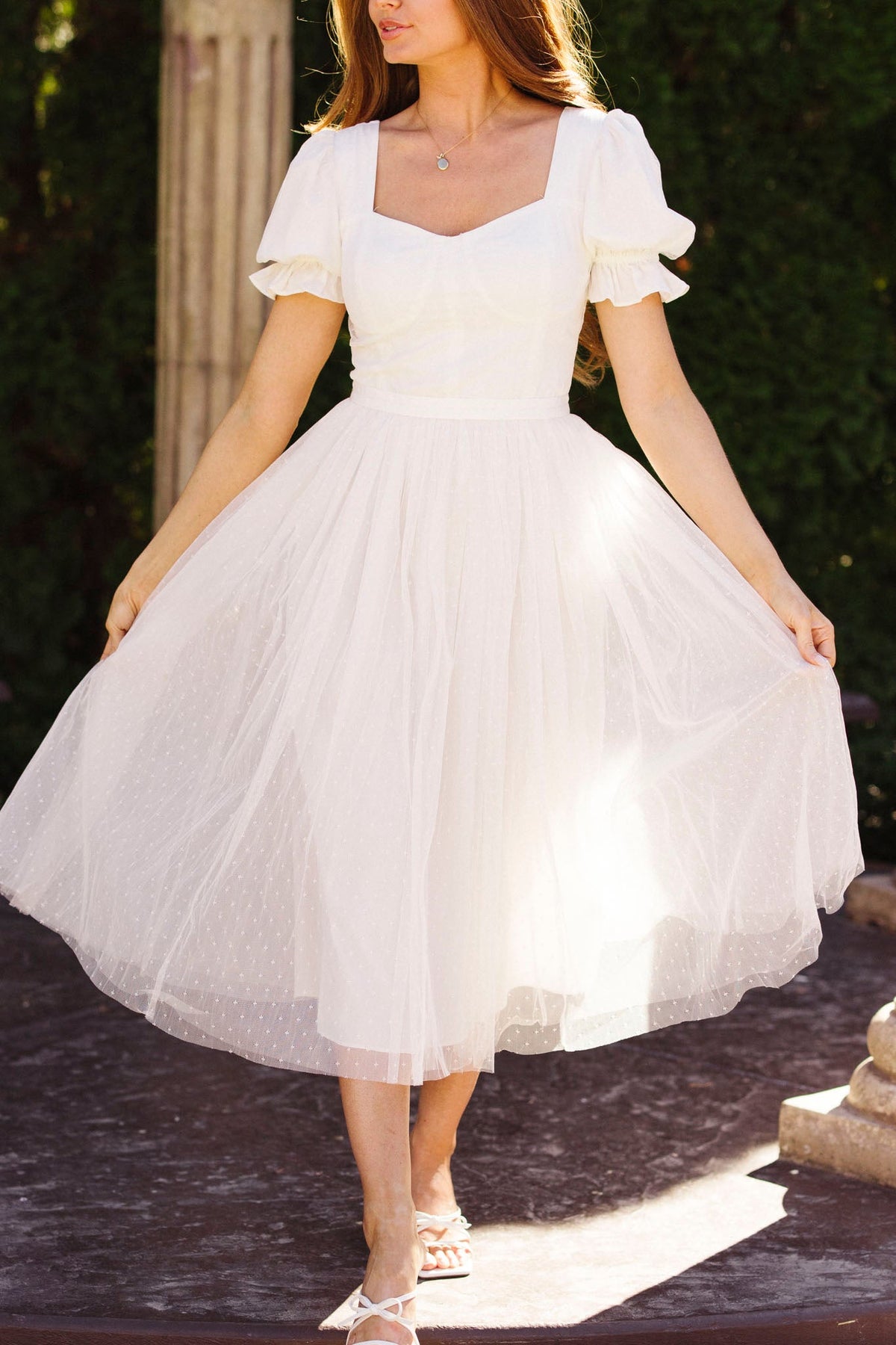 A woman wears the Ivy City Co Ballerina Dress in White, featuring a fitted bodice and flared tulle skirt. The light, short-sleeved style adds charm as she stands outdoors, holding the sides of her dress with her face partly out of frame.