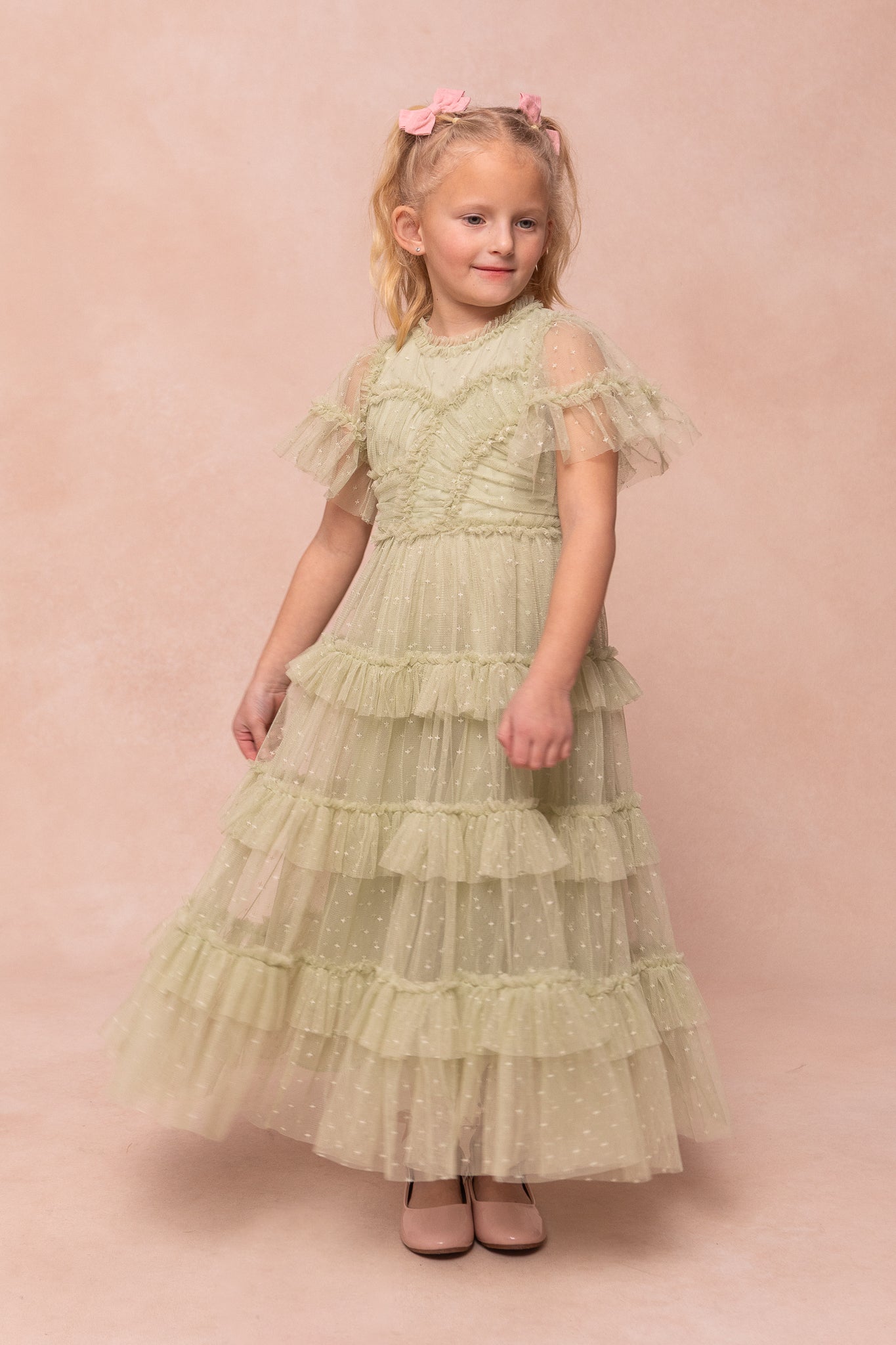 A young blonde girl with pink bows wears the Ivy City Co Mini Whimsical Maxi Dress in Sage, featuring flutter sleeves and sheer dotted fabric, standing on a pale pink background and smiling slightly as she twirls.