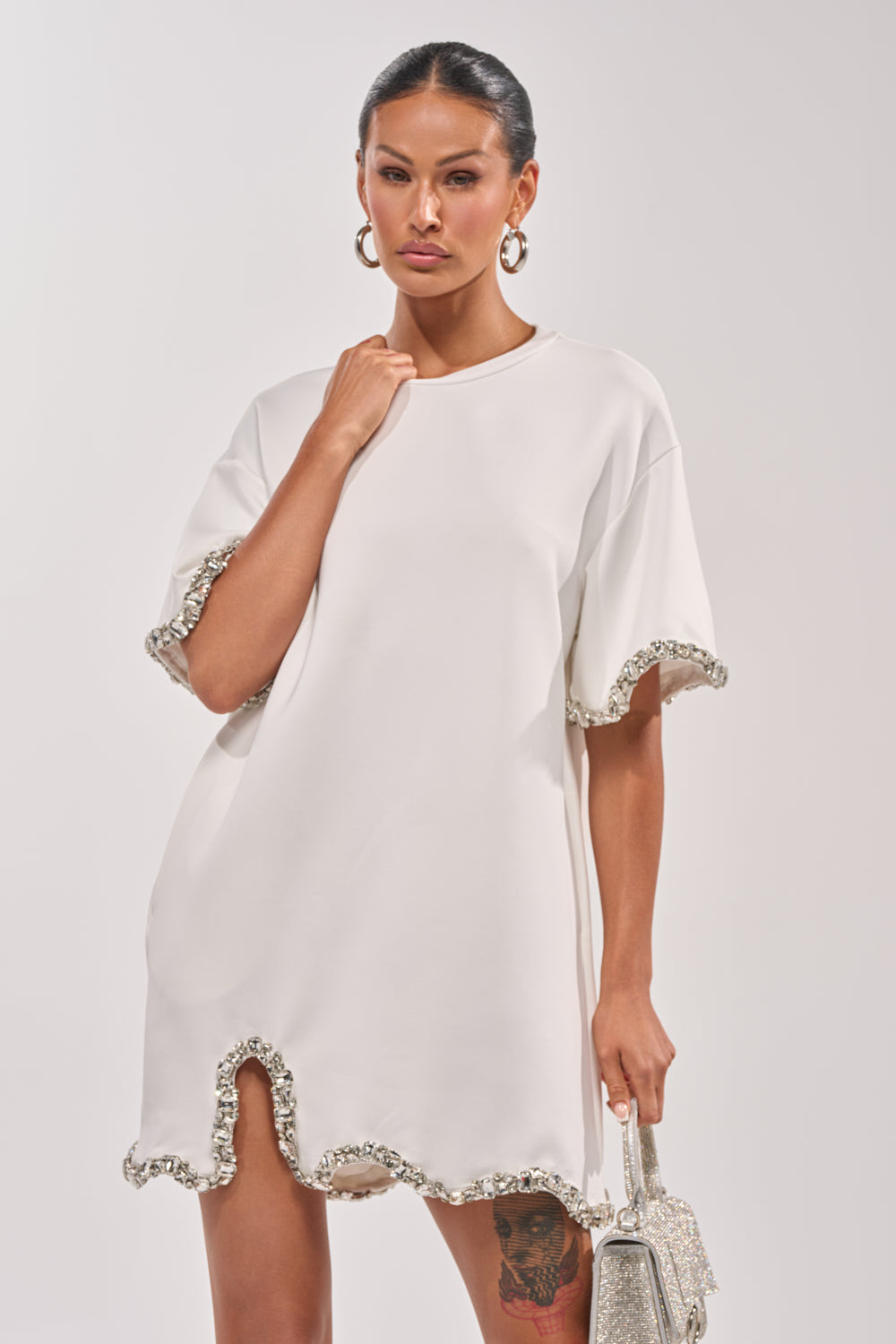 A woman in the COOL GIRL EMBELLISHED TEE SHIRT DRESS IN WHITE with jeweled trim and sleeves holds a sparkly handbag. She has hoop earrings, a sleek hairstyle, and a visible tattoo on her left thigh.