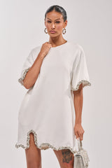 A woman in the COOL GIRL EMBELLISHED TEE SHIRT DRESS IN WHITE with jeweled trim and sleeves holds a sparkly handbag. She has hoop earrings, a sleek hairstyle, and a visible tattoo on her left thigh.