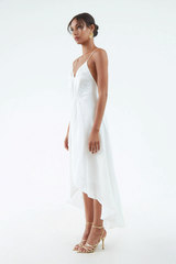 Side view of a white silk-hemp dress with thin shoulder straps, V-neck line, a gathered waist, and a flared hem that's short in the front and long in the back. Model is wearing gold sandals and earrings. 