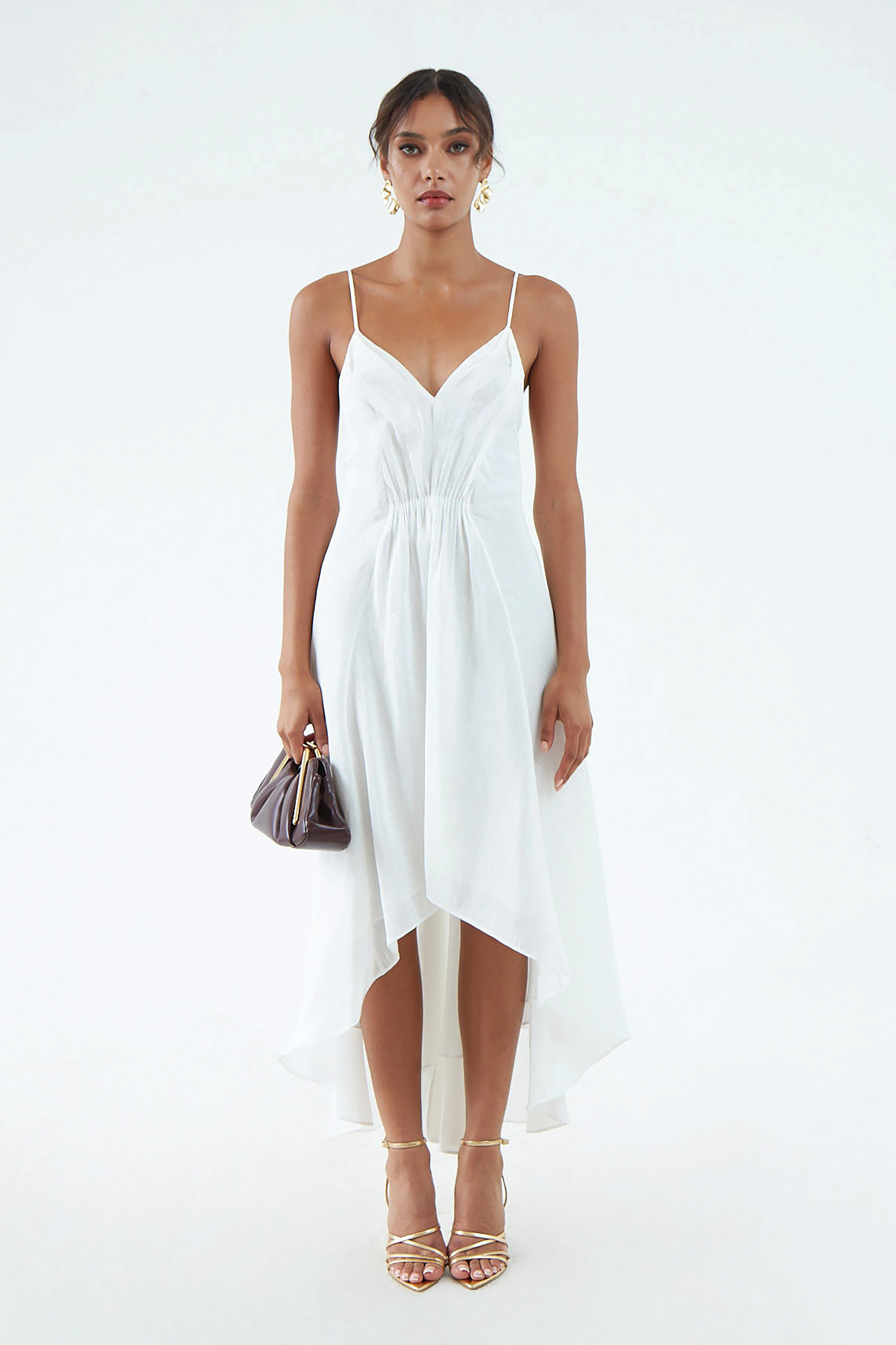 White silk-hemp dress with thin shoulder straps, V neckline, waist gather, and flared, asymmetrical hem that waterfalls around the legs. Worn with gold sandals and maroon clutch.