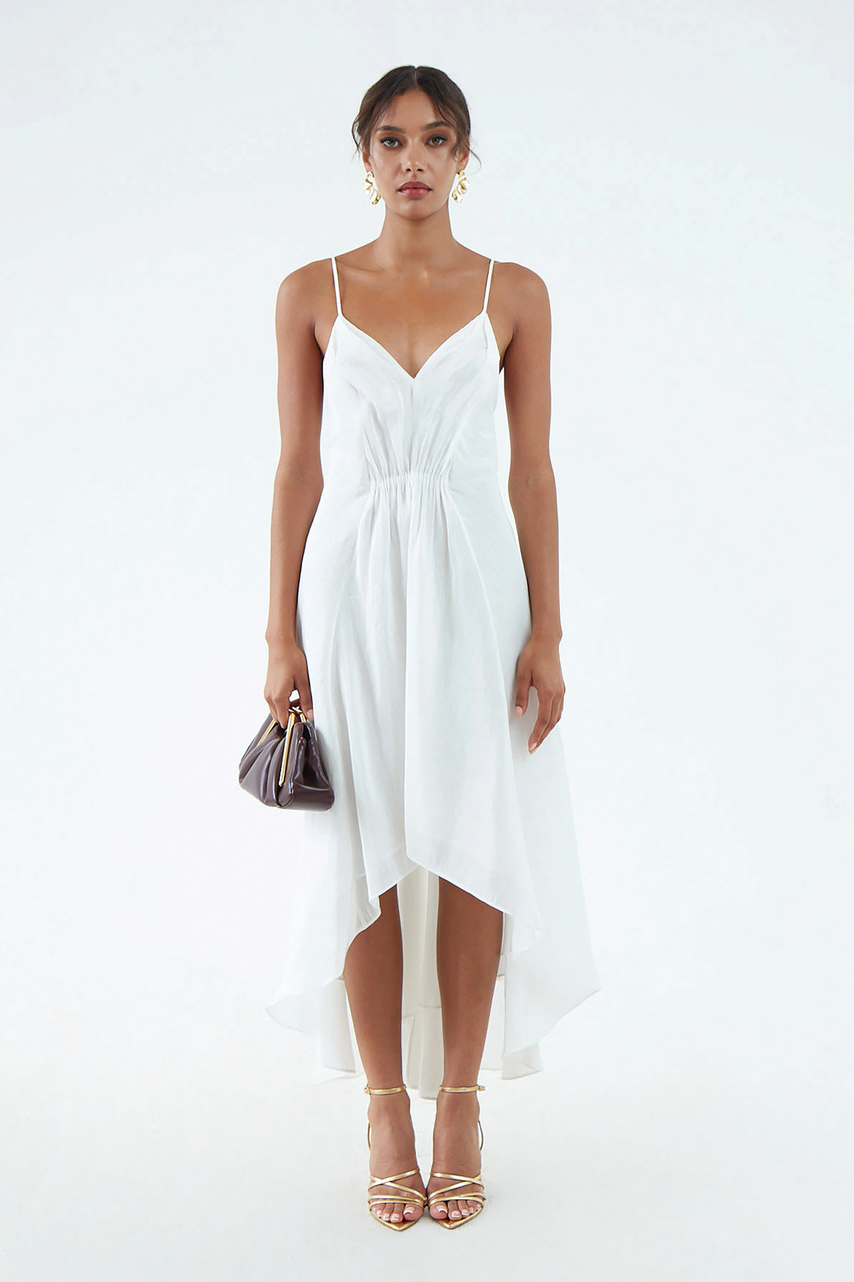White silk-hemp dress with thin shoulder straps, V neckline, waist gather, and flared, asymmetrical hem that waterfalls around the legs. Worn with gold sandals and maroon clutch.