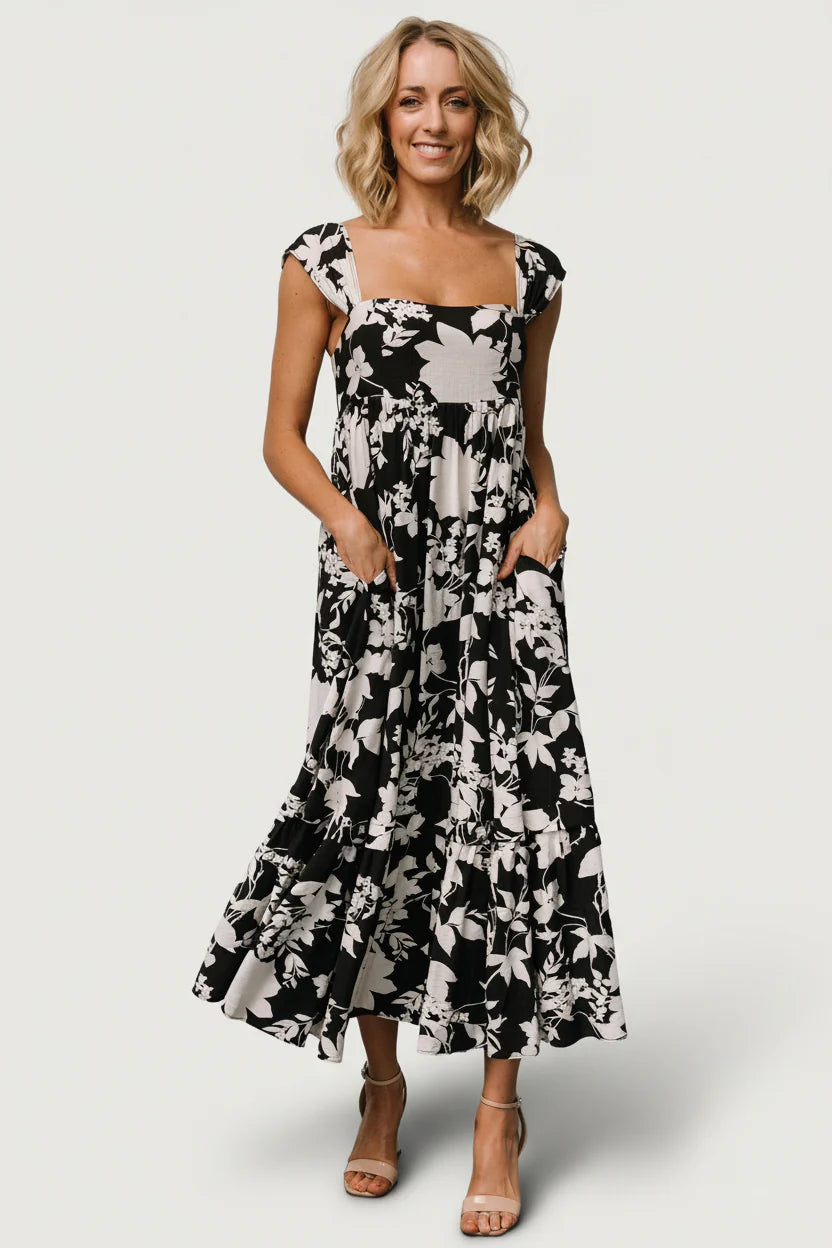 Black and ivory floral maxi dress on model, showing square neckline, gathered cap sleeves, smocked back and flowing single-tiered skirt with side pockets.