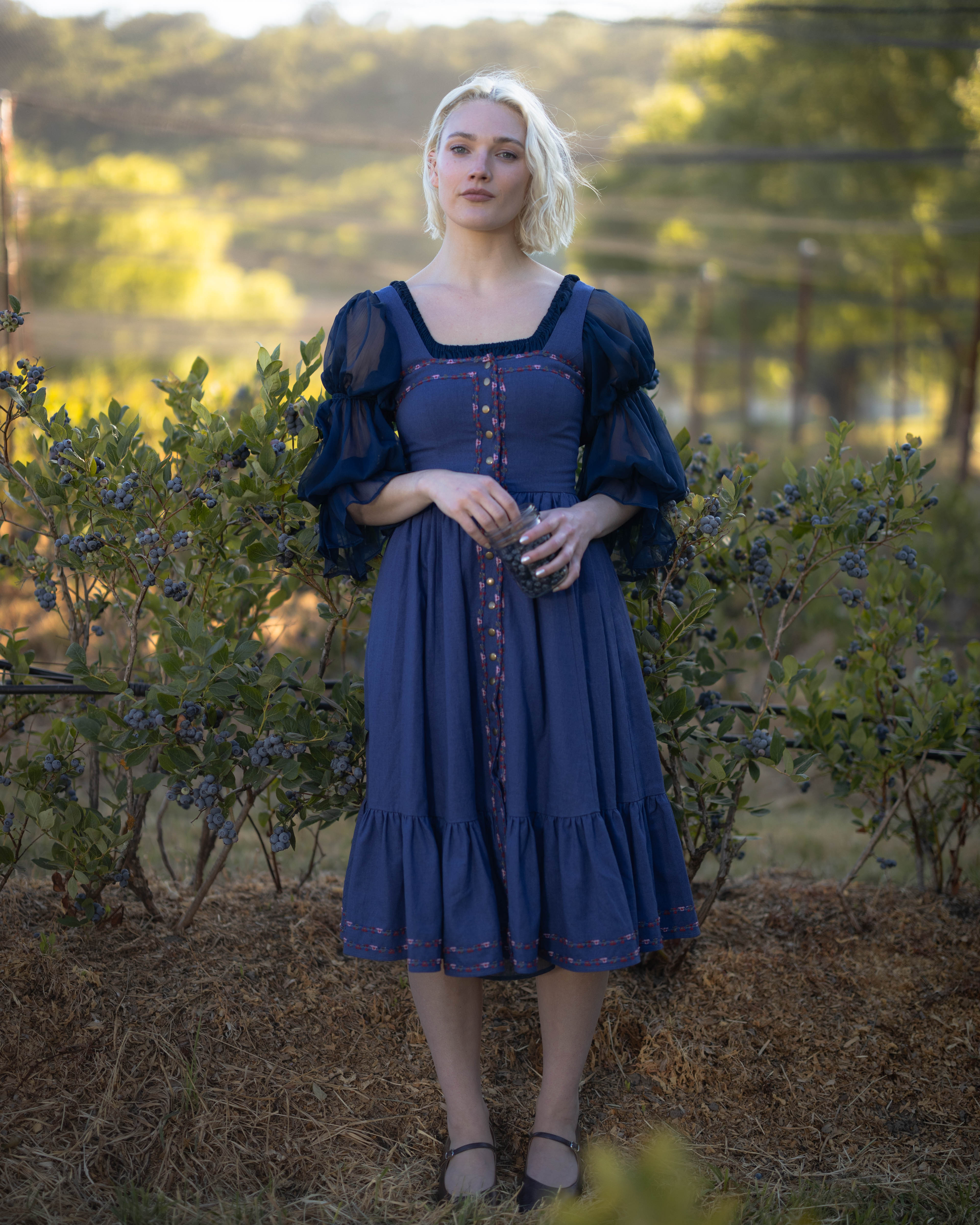 Woman wearing navy blue chiffon chemise with puff sleeves standing outdoors near bushes in soft sunlight