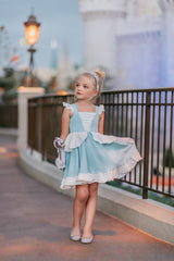 A young girl in an "Our Original Cinderella Twirl Dress" stands near a castle.  The dress has a white flutter ruffle on the straps. There is a white skirt ruffle that overlays the blue twirl skirt. The soft cotton material is comfortable and breathable.