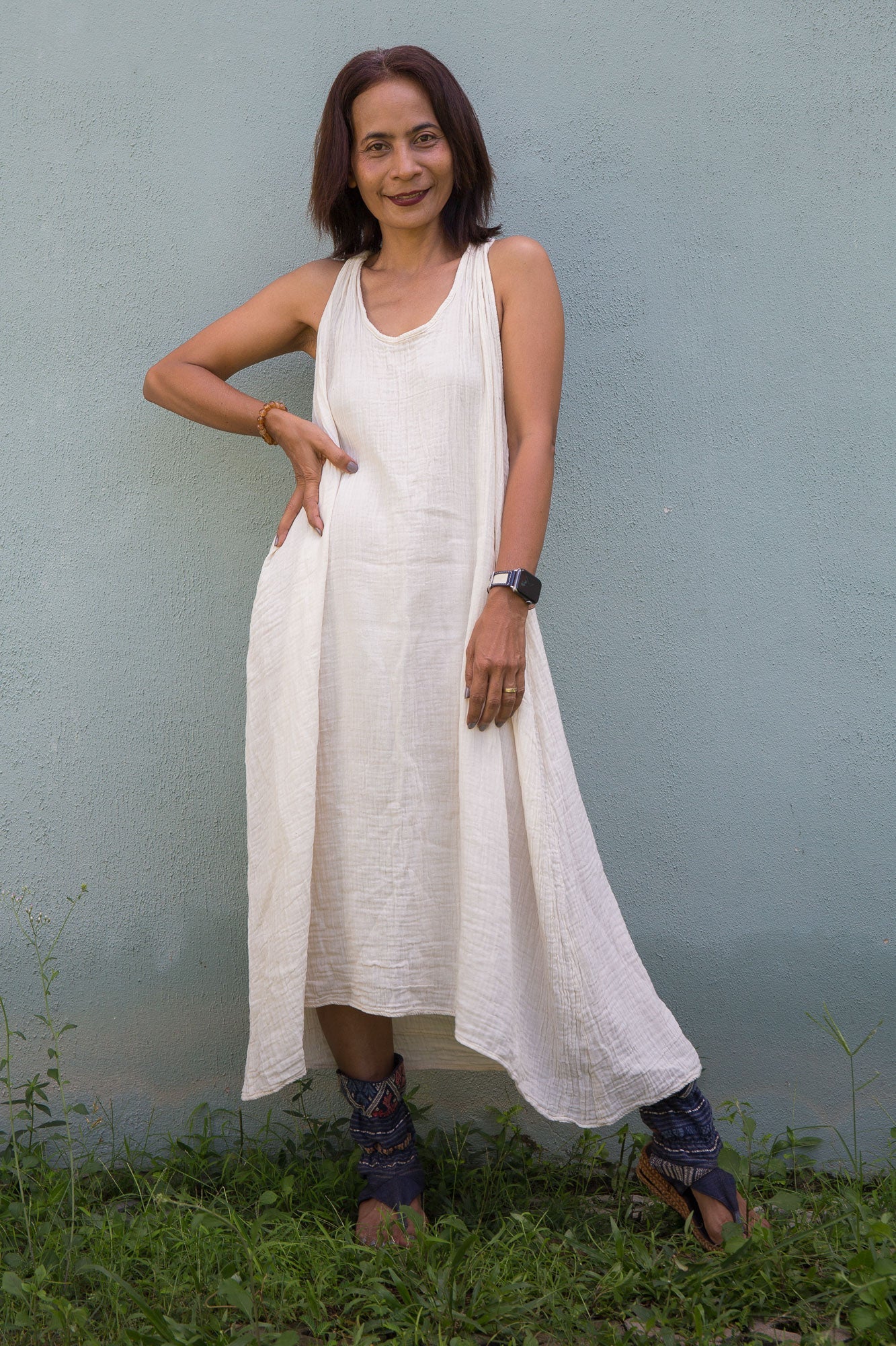 Woman wearing a white cotton gauze dress standing against a light blue wall with green grass at the bottom.