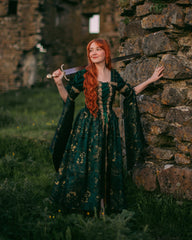 Woman in medieval ruins wearing a Dark Green Jacquard Corset Dress with gold accents and removable bell sleeves.
