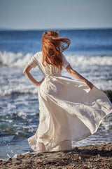 Woman wearing creamy summer linen wedding dress standing by the ocean shore with flowing skirt and tie wrap waist