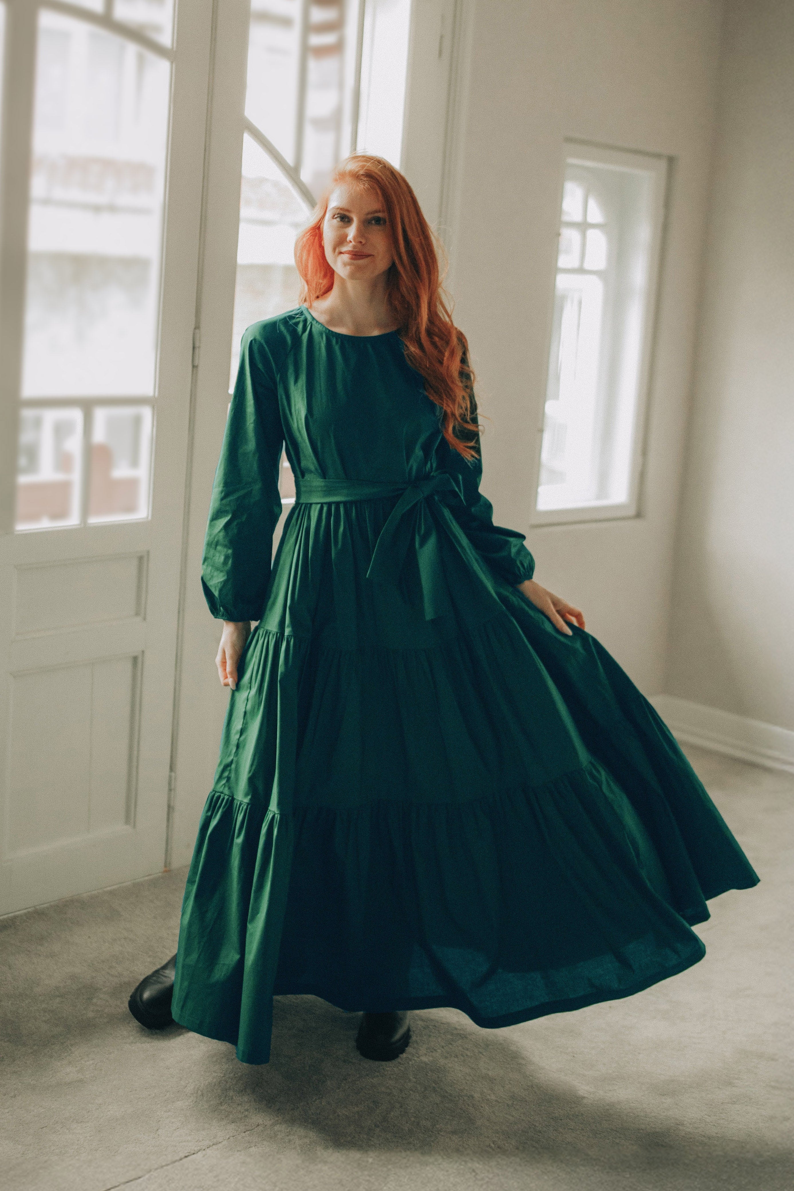 Woman wearing a dark green tiered cotton maxi dress with long sleeves and a tied waist in a bright room