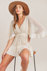 Cream lace V-neck mini romper with wide sleeves and button-down front, paired with a brown hat, seated pose.
