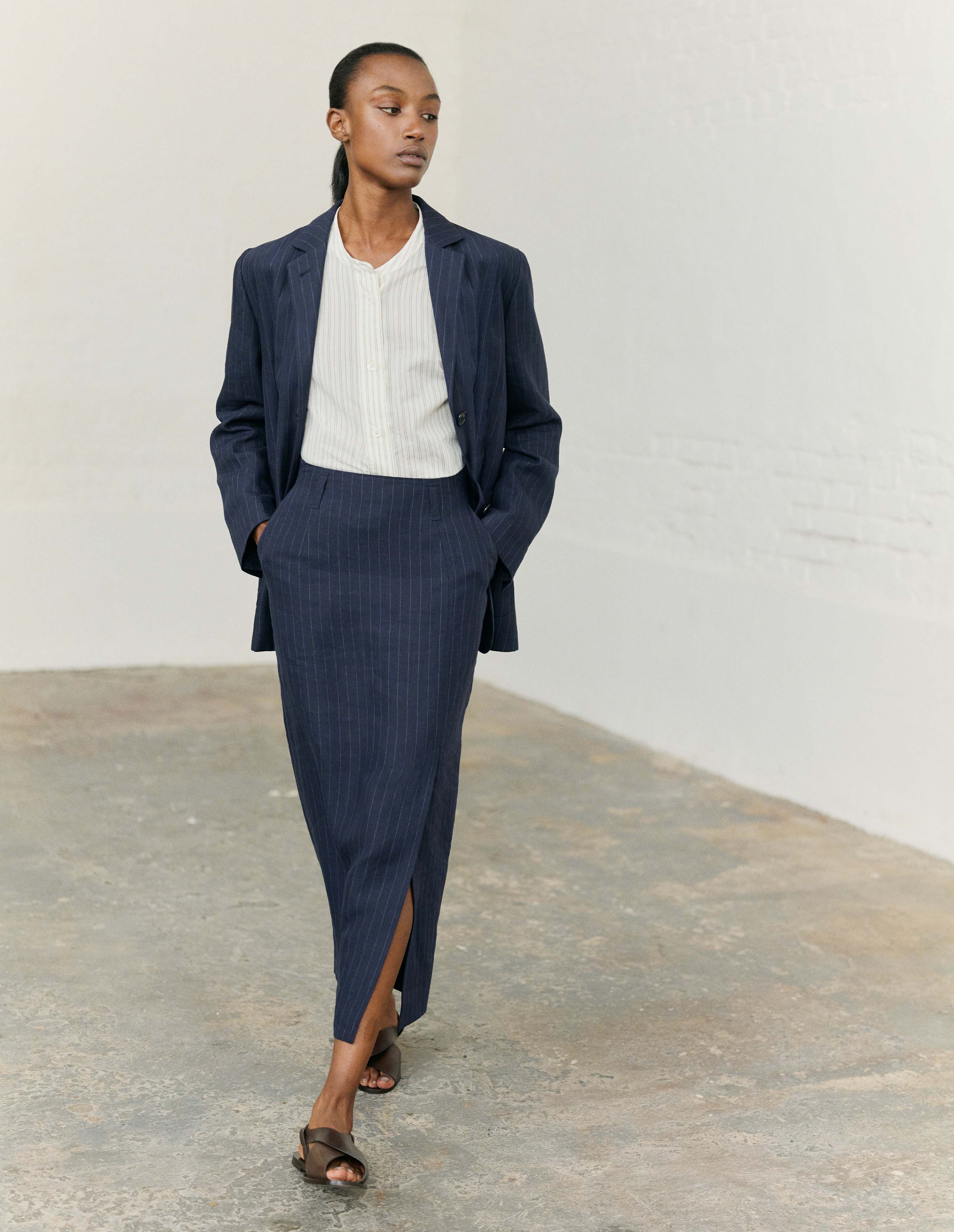 Full body image, front view of model wearing navy and grey pinstripe linen skirt, with offset front vent, slant side pockets and belt loops. Styled with matching navy and grey pinstripe blazer and collarless white and grey pinstripe shirt with dark brown leather sandals.