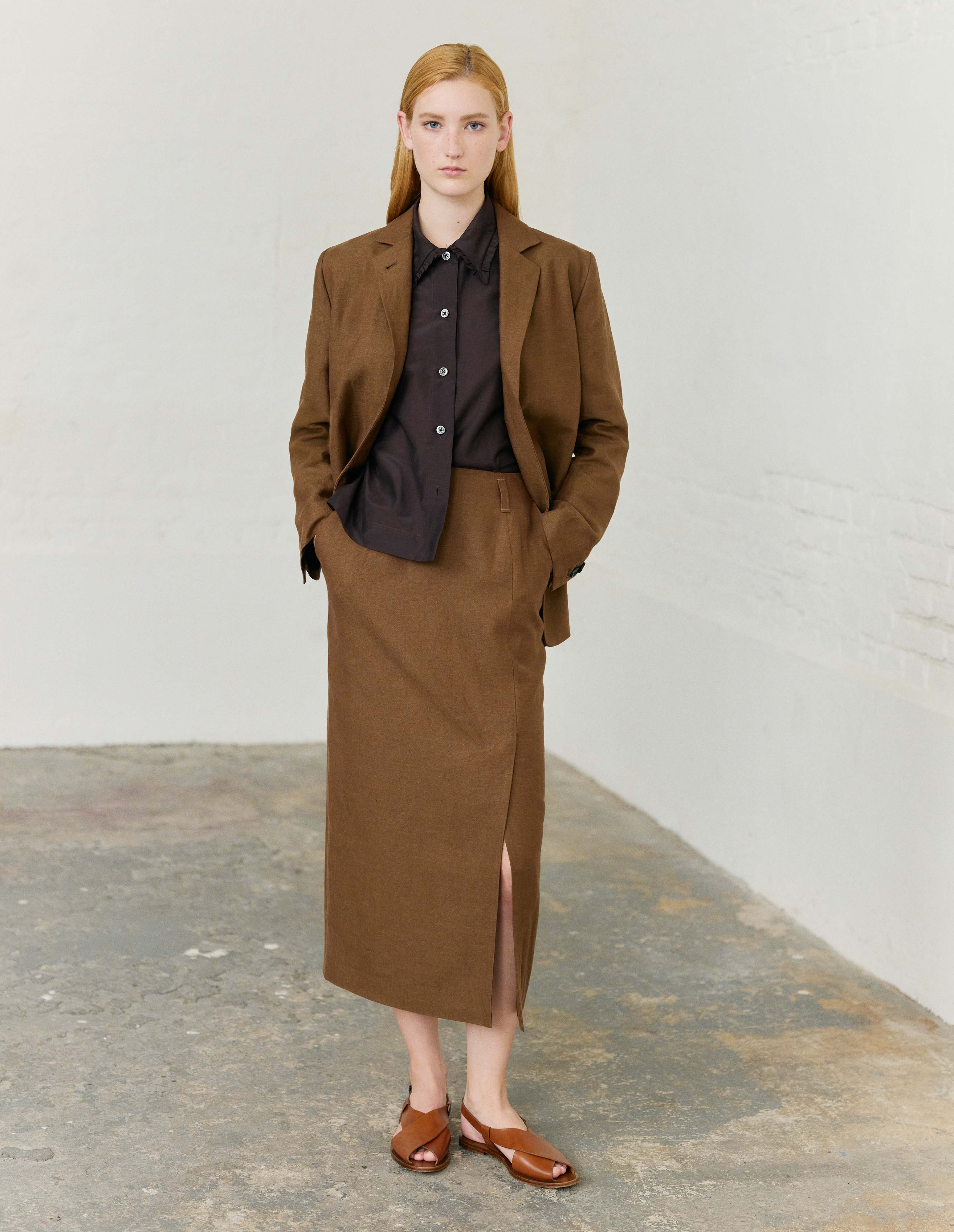 Full body image, front view of model wearing brown linen skirt, with offset front vent, slant side pockets and belt loops. Styled with matching brown blazer, dark brown ruffle collared shirt and light brown leather sandals.