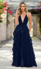 A woman with long blonde hair wears the Velvi Ruffled Long A-Line Prom Dress Alances, a sleeveless deep V-neck navy gown, while standing outdoors on a flower-lined pathway.