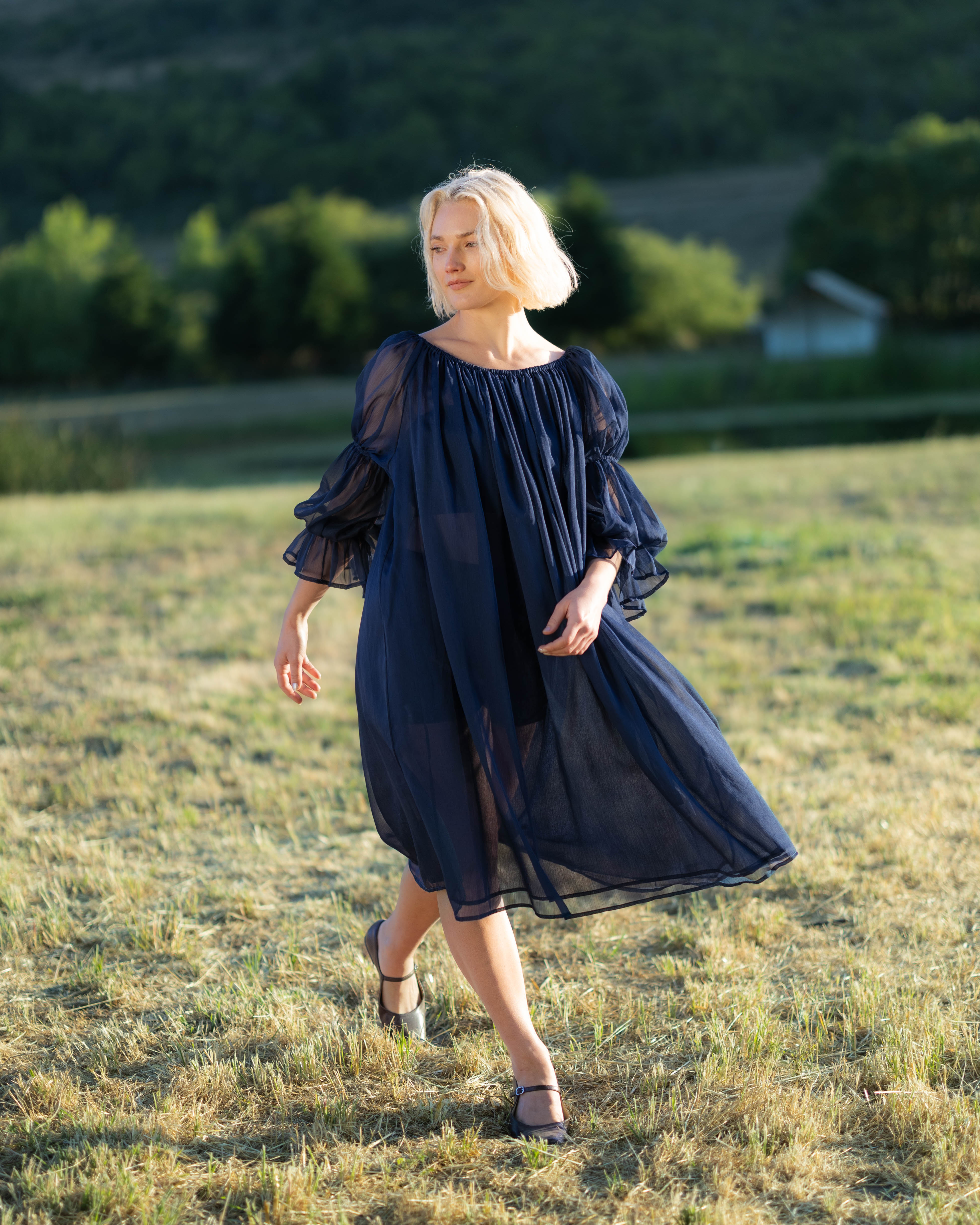 Woman wearing a flowing navy blue chiffon chemise with puff sleeves walking outdoors in a grassy field