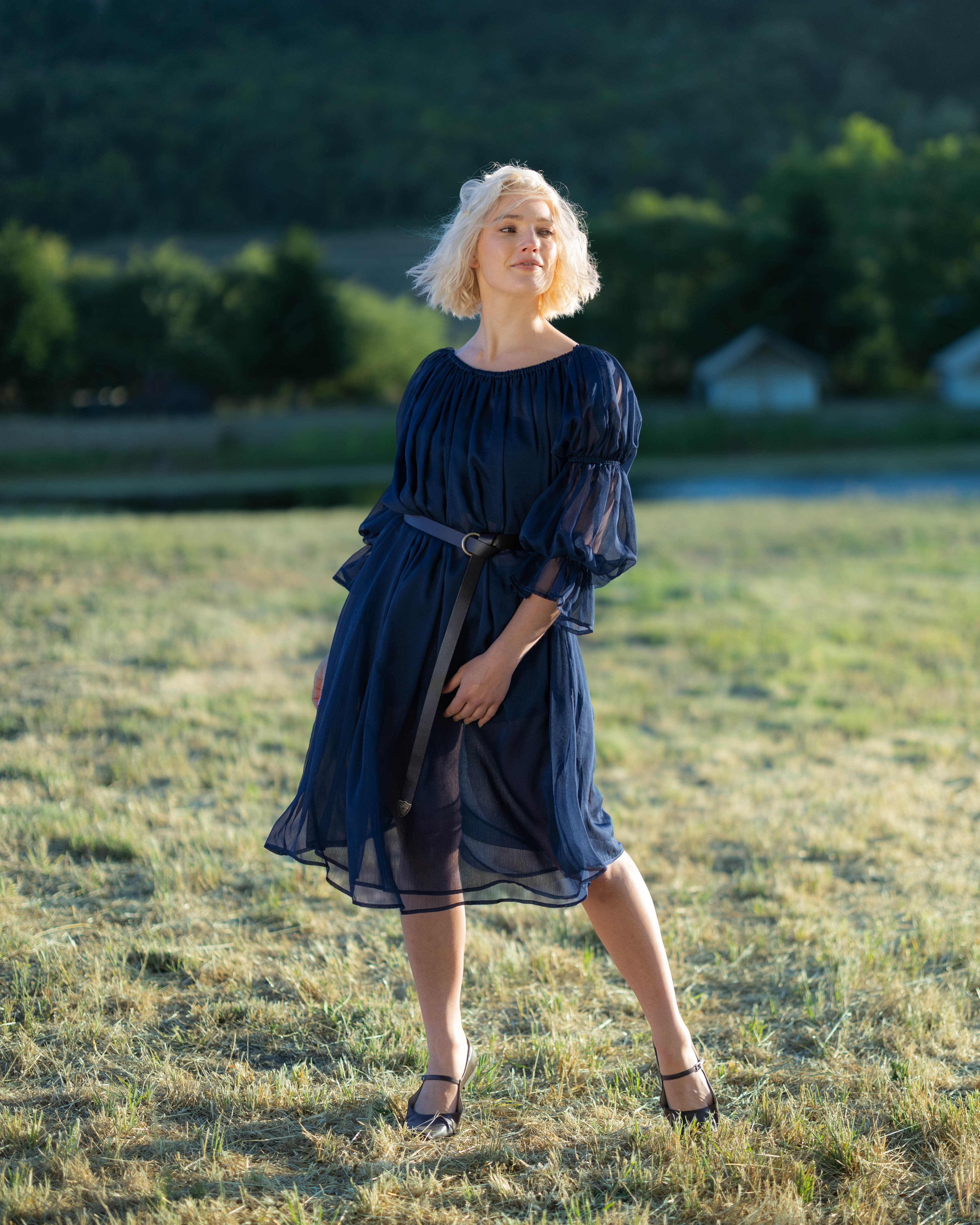 Woman wearing navy blue chiffon chemise with puff sleeves and belt, standing outdoors on grassy field at sunset