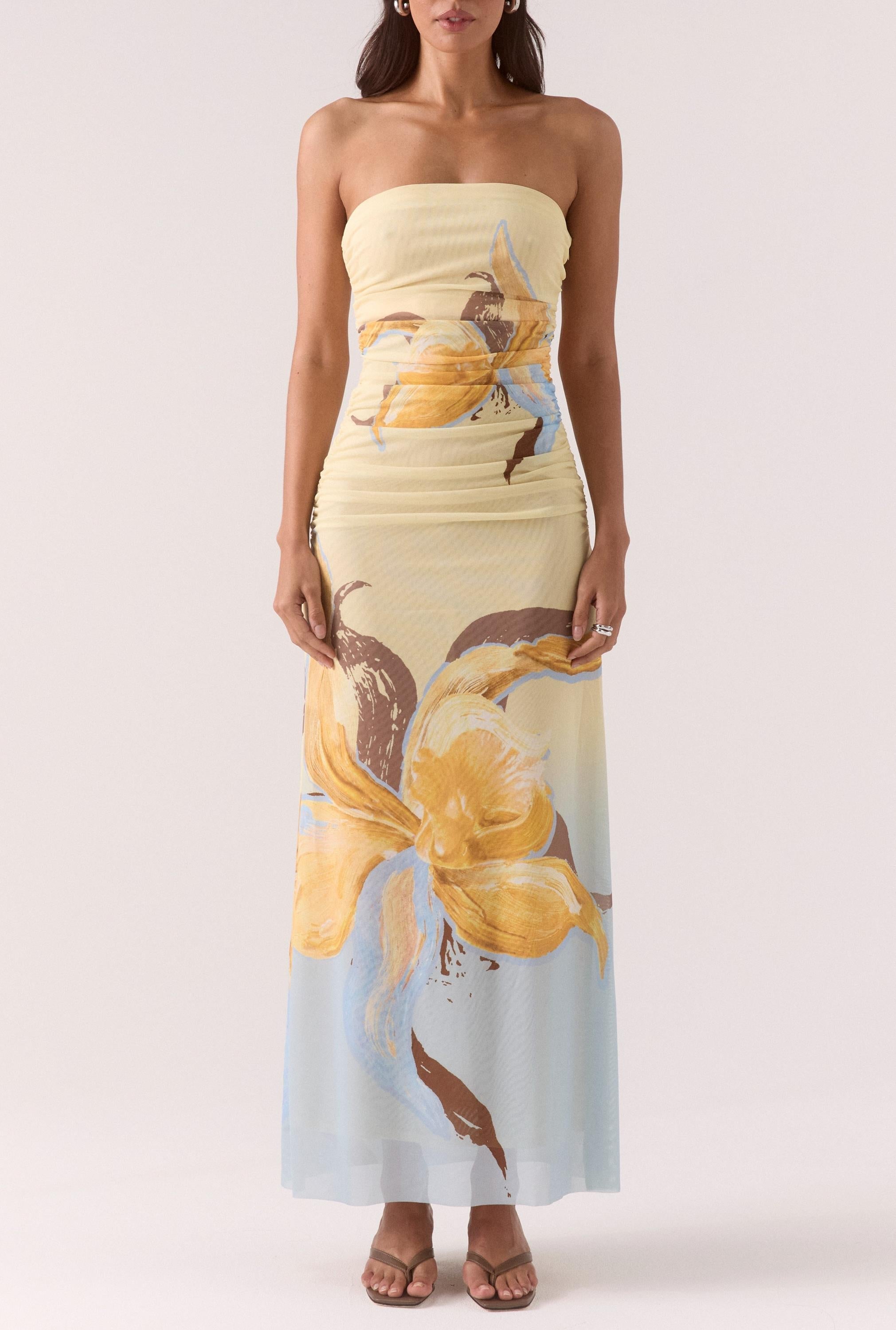 A woman stands with arms by her sides, wearing the Sovere Dahlia Maxi Dress—a strapless stretch mesh maxi in large yellow and brown florals over a light pastel background—paired with open-toe sandals.