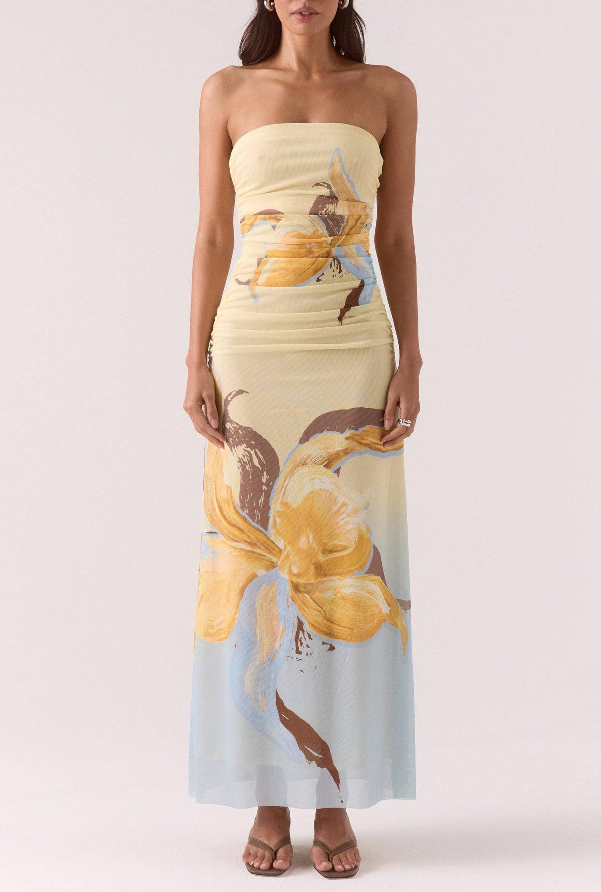 A woman stands with arms by her sides, wearing the Sovere Dahlia Maxi Dress—a strapless stretch mesh maxi in large yellow and brown florals over a light pastel background—paired with open-toe sandals.