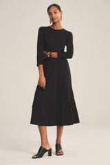 A woman models the RIVER DRESS by Velvet by Graham & Spencer, a long-sleeve black cotton slub A-line dress, styled with black heeled sandals, red earrings, and bracelets, standing against a plain light background.