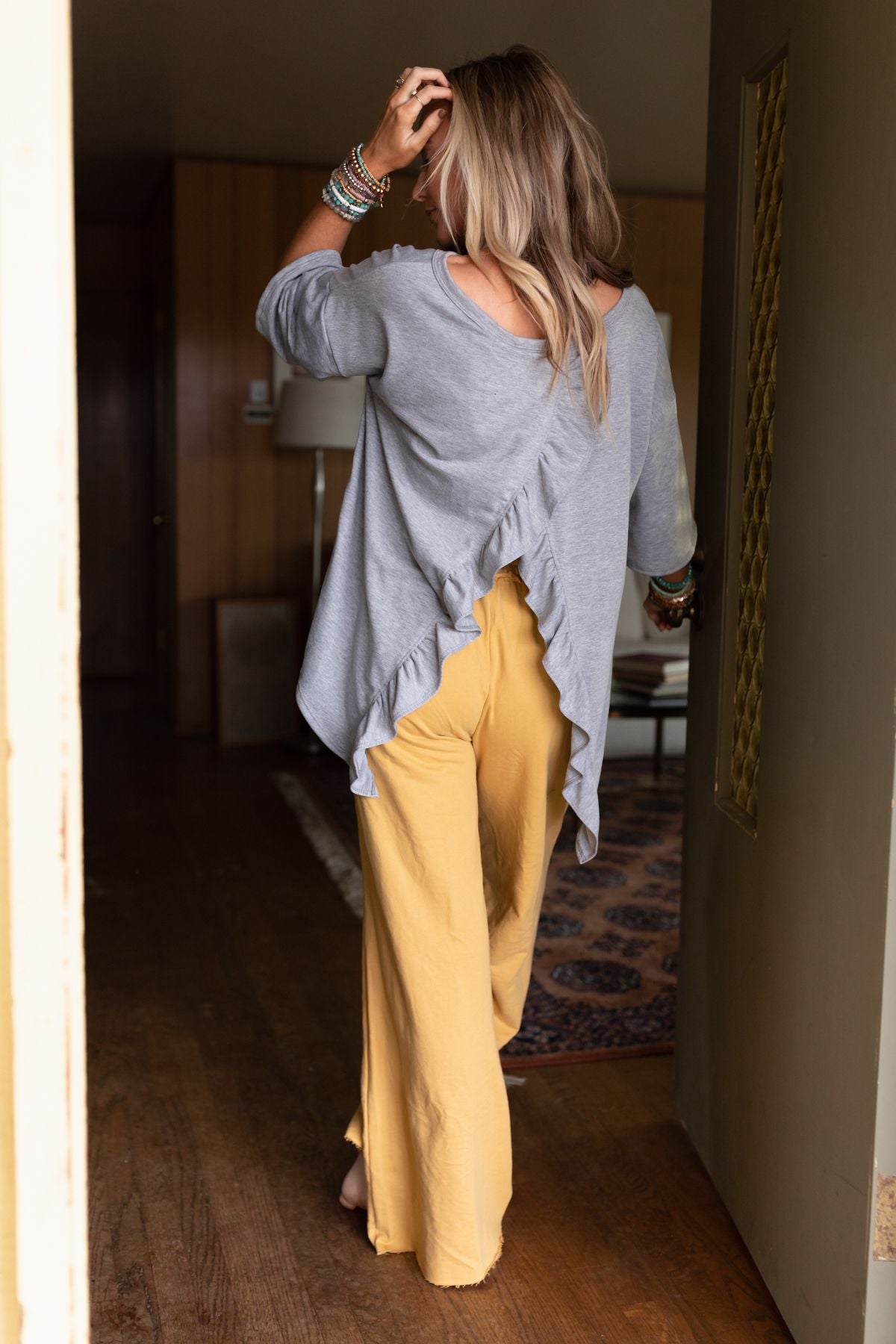 A woman with long hair, barefoot and facing away, walks in a wooden room wearing The Nest Must Have High Low Tee in Heather Gray with wide-legged yellow pants.