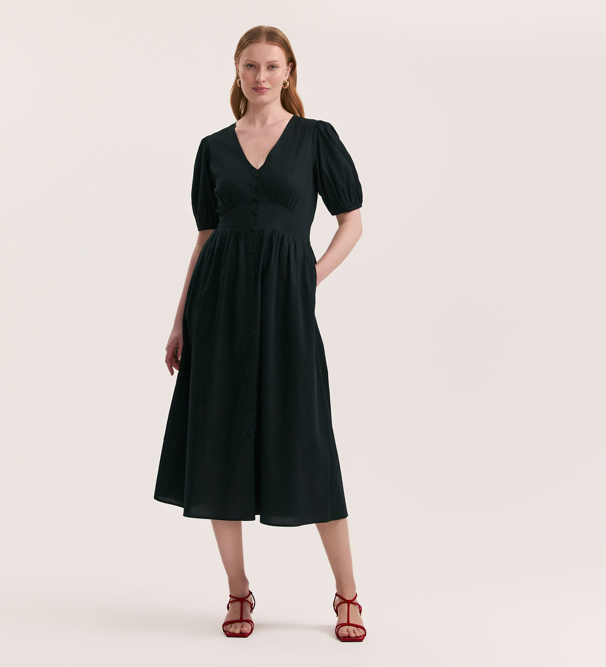 A woman stands against a plain background wearing the Addison Black Cotton Midi Tea Dress, featuring a v-neck, short puff sleeves, and a fitted waist, paired with red strappy heels.