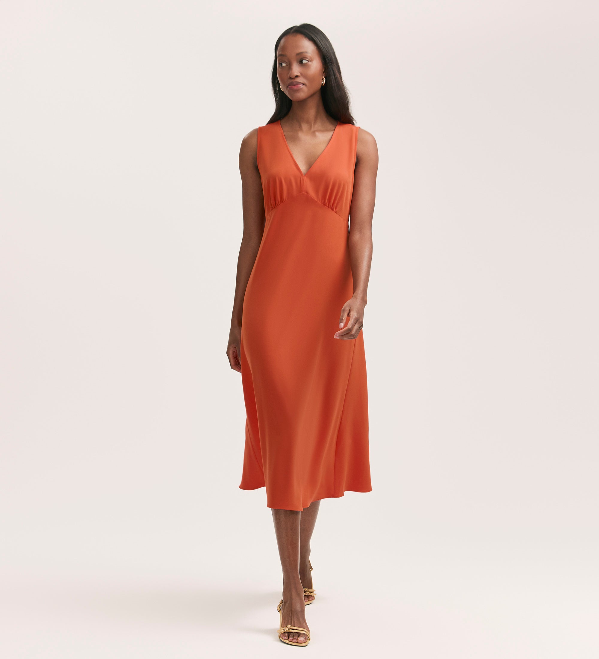 A woman models the Brionie Orange Crepe Sleeveless Midi Dress, featuring crease-free fabric, standing against a plain light background with a relaxed expression.
