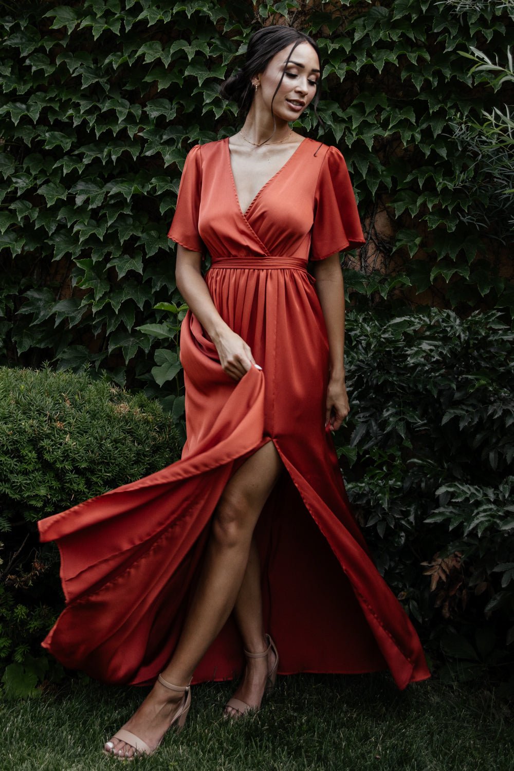 Full-length front view of rust satin-like maxi dress with surplice V-neck, flutter sleeves, elastic waist and removable sash worn by a model.