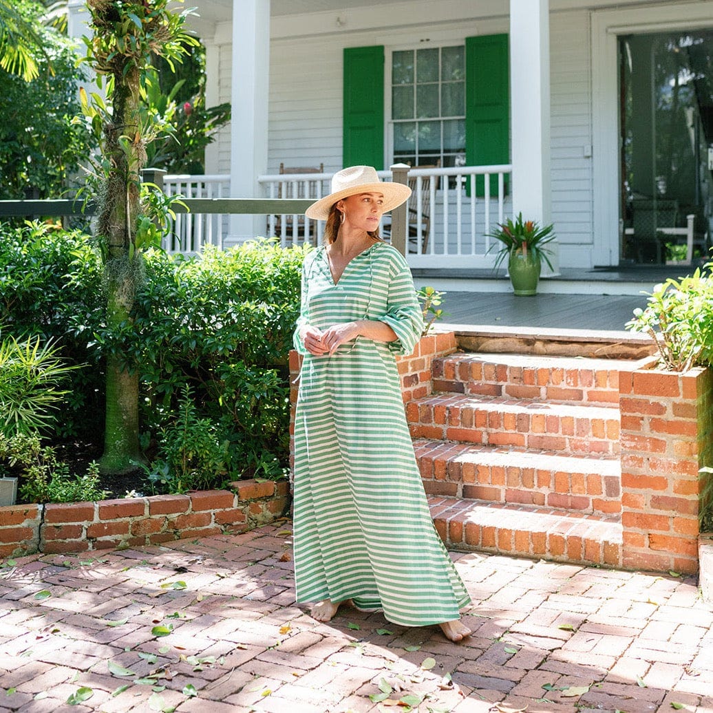 A woman standing on a brick pathway wearing a green and white striped kaftan dress with a v-neck, sleeveless, and a side slit. She is also wearing a wide-brimmed hat.