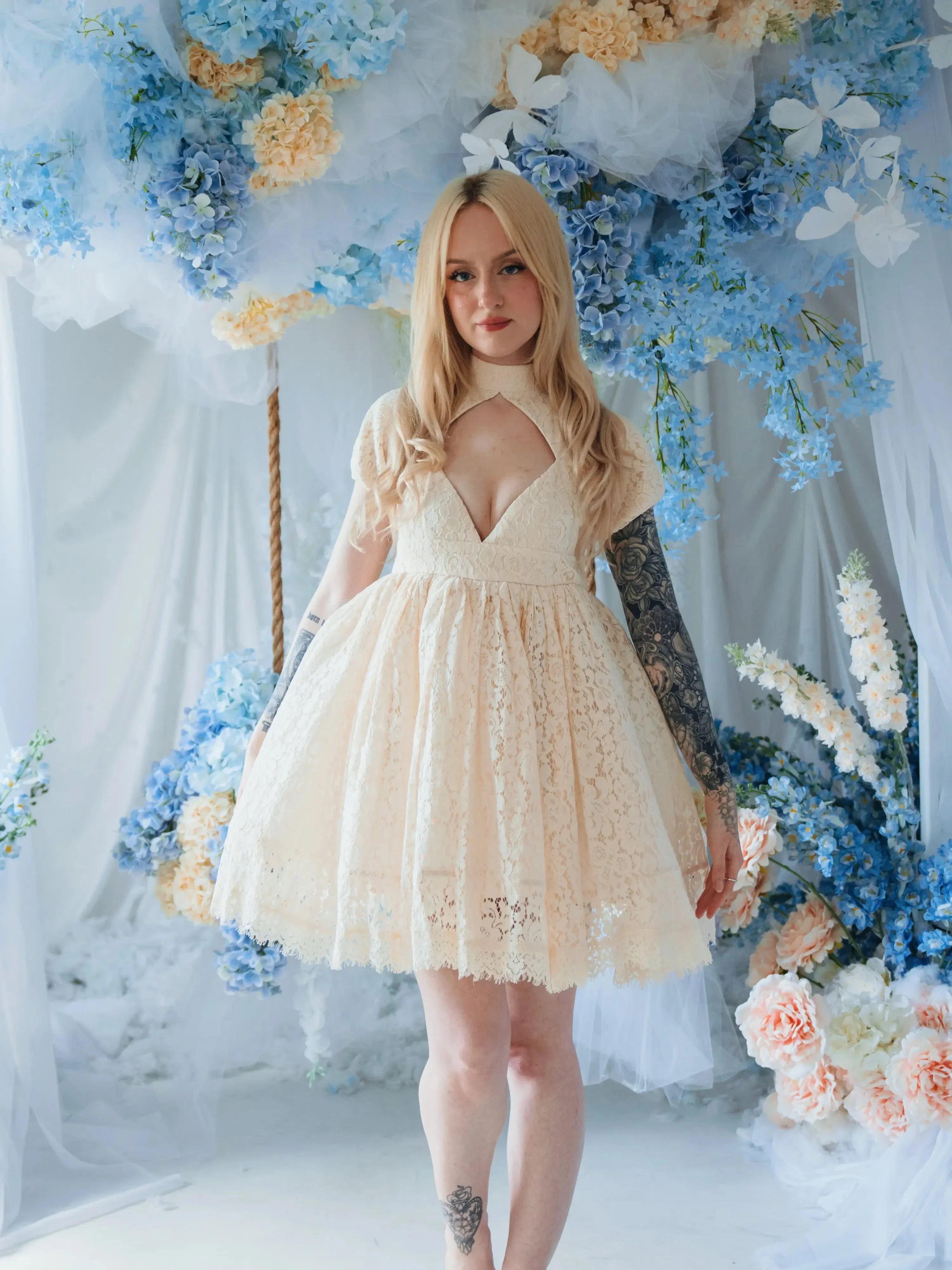 The Victorian Lace Tea Party Mini Dress in off white lace with puff sleeves on a model and styled in a floral setting.