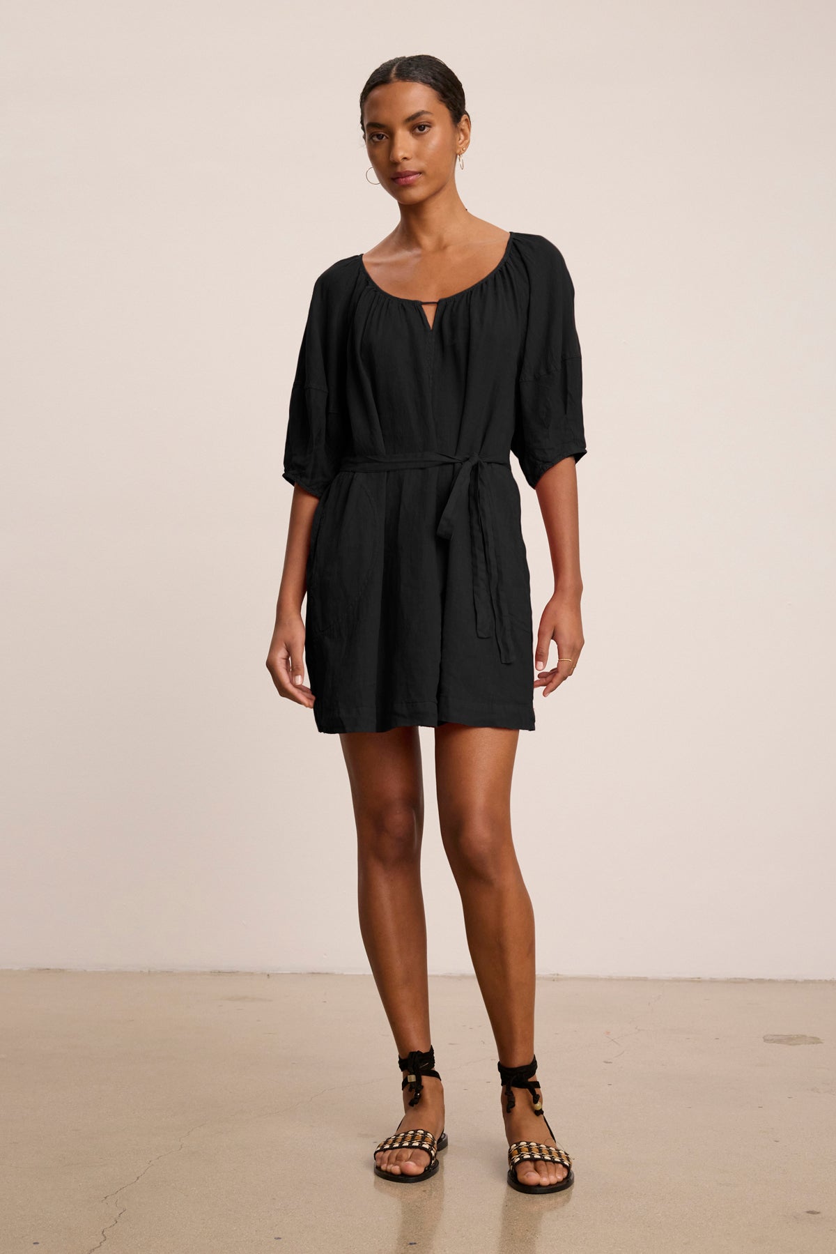 A woman stands indoors against a plain beige background wearing the GRETA LINEN DRESS by Velvet by Graham & Spencer—a black, short-sleeved, belted woven linen mini dress—paired with black sandals.