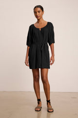 A woman stands indoors against a plain beige background wearing the GRETA LINEN DRESS by Velvet by Graham & Spencer—a black, short-sleeved, belted woven linen mini dress—paired with black sandals.