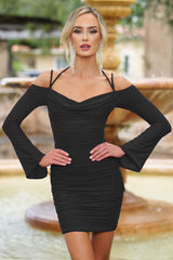 A woman stands confidently in a fitted black dress with long, flared sleeves. She poses with her hands on her hips, set against a decorative stone fountain in a garden.